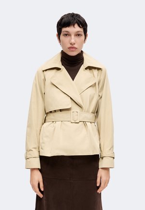 Personne aux cheveux courts et foncés portant un trench-coat beige ceinturé par-dessus un col roulé brun foncé et une jupe assortie, debout devant un fond uni.