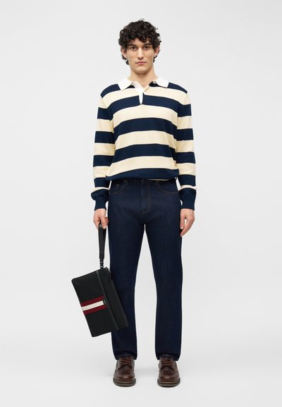 Jeune homme portant un maillot de rugby rayé bleu marine et blanc, un jean foncé et des chaussures marron, tenant un sac noir avec des rayures rouges et blanches.