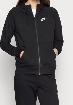 Kvinde iført sort Nike hættetrøje med lynlås og lommer samt matchende sorte Nike bukser, hænderne i hættetrøjelommerne.