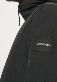 Svart lättviktsjacka med en texturerad yta, med huva och en rektangulär tygpatch med "Calvin Klein"-logotyp på ärmen.