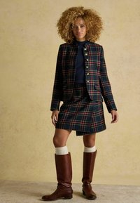 Kariertes Woll-Blazer mit goldenen Knöpfen, kombiniert mit einem passenden karierten Rock. Dazu gehören knielange braune Lederstiefel und weiße Kniesocken.