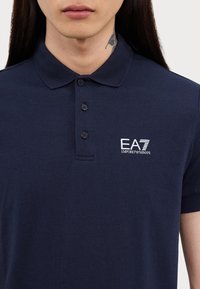 Námornícka polokošeľa vyrobená z bavlny, s klasickým golierom, tromi gombíkmi a bielym vyšitým logom EA7 na ľavej hrudi.