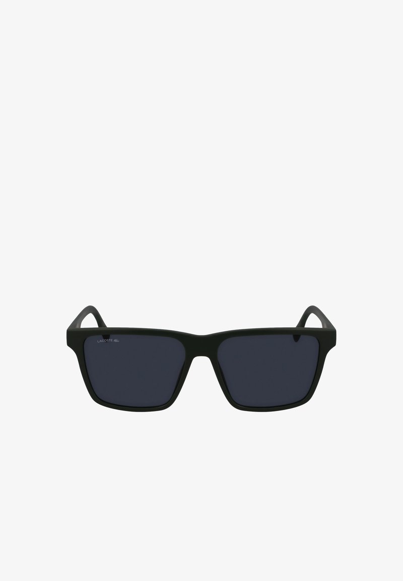 Mustat suorakaiteen muotoiset aurinkolasit tummilla linsseillä, kumipintainen matta kehys ja hillitty logo vasemmassa linssissä. Minimalistinen muotoilu.