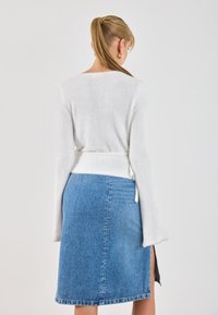 Weißer flauschiger Pullover mit einer Schleife an der Taille, kombiniert mit einem hellblauen Jeansrock mit einem seitlichen Schlitz und einer Saumstickerei.