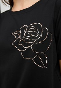 Melns T-krekls ar rozes dizainu no rozā zelta rhinestones, ar gludu auduma tekstūru un klasisku apaļu kakla izgriezumu.