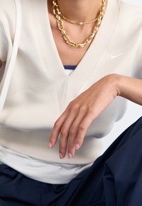 Ljusbeige v-ringad topp med ribbad struktur och korta ärmar. Ett guldkedjehalsband lager på lager över en blå topp som syns under.