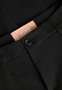 Zwarte stoffen broek met een gladde textuur, voorzien van een zwarte knoop en een licht perzikkleurig label met "JJXX" in het wit.
