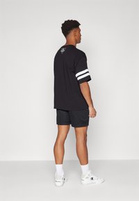 Camiseta negra de gran tamaño con rayas blancas en las mangas, combinada con pantalones cortos de malla negros. El modelo lleva zapatillas blancas y calcetines.