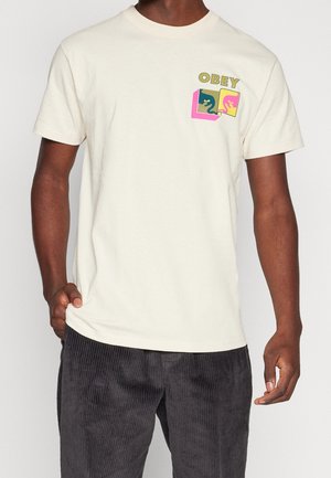 Kremowa bawełniana koszulka z grafiką na kieszeni piersiowej z napisem "OBEY", kolorowymi postaciami z kreskówek oraz jaskrawymi akcentami geometrycznymi.