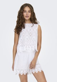Top bianco corto con ricami a giorno e orlo a scollatura, abbinato a pantaloni corti bianchi coordinati con vita regolabile e bordi decorativi.
