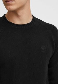 Black knitted sweater with a crew neck, featuring ribbed details at the neckline and cuffs, and a small logo on the chest.