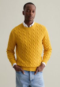 Gelber Kabelstrickpullover mit gerippten Bündchen und Saum, getragen über einem weißen Hemd mit Kragen, ergänzt durch hellblaue Jeans.