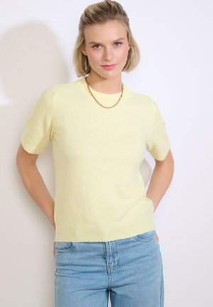 Junge Frau mit kurzen blonden Haaren, die einen hellgelben Kurzarmpullover, hellblaue Jeans und eine goldene Kettenhalskette trägt und mit den Händen am Rücken posiert.