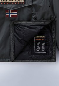 Vesi- ja tuulenpitävän tummanvihreän takin sisäosa, jossa on eristys, Norjan lipun merkki ja hoitotarra, jossa on vedenpitävät ja hengittävät ikonit.