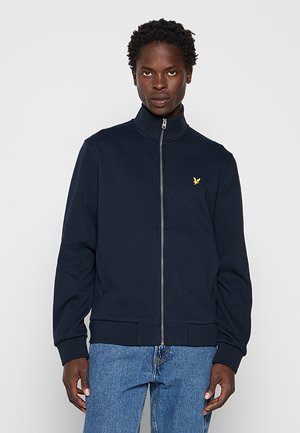 Man draagt een marineblauwe jas met rits en een geel vogel-logo op de borst en blauwe spijkerbroek, staand tegen een effen lichte achtergrond.