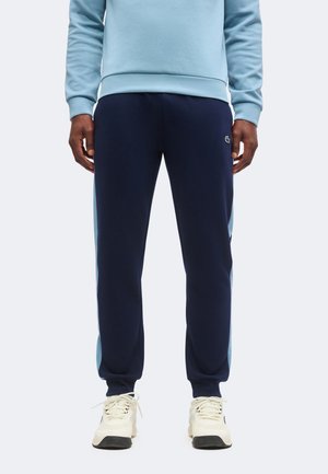 Uomo che indossa pantaloni della tuta blu navy con pannelli laterali azzurri, una felpa azzurra e sneakers crema, in piedi su sfondo semplice.