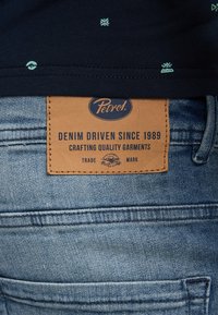 Blauwe spijkerbroek van denim met een tan lederen label waarop met blauwe tekst "Petrol" staat, met de details "Denim Driven Since 1989" en "Crafting Quality Garments".