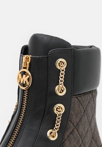 Gros plan sur une botte en cuir noir et tissu matelassé avec une fermeture éclair dorée et un détail de chaîne, ornée d'un curseur de fermeture éclair avec le logo MK.