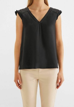 Femme portant un chemisier noir sans manches en V avec des détails en dentelle et un pantalon beige, debout devant un fond blanc uni.