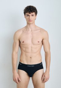 Schwarze Baumwollunterhose mit einem kontrastierenden weißen Calvin Klein-Logo am Bund, Modell mit durchtrainierter Figur, steht vor einem hellen Hintergrund.