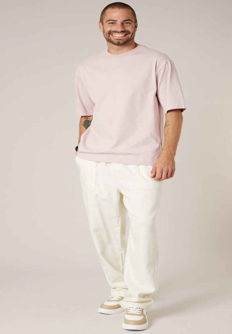 Homme souriant debout avec les mains dans les poches, portant une chemise à manches courtes rose pâle, un pantalon blanc et des baskets beige et blanches.