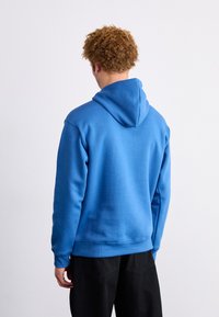 Moletom azul feito de um tecido suave e texturizado, apresentando um capô com cordão e punhos canelados. Calças pretas também são visíveis abaixo da cintura.