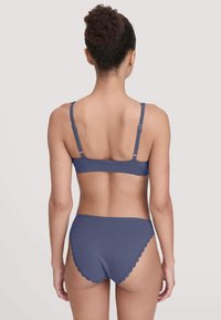 Vrouw met donker gekruld haar in een knot, gekleed in een bijpassende blauwe bh en hoge stijlen onderbroek, gezien van achteren op een effen achtergrond.