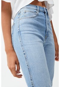Jean taille haute bleu clair en denim, avec une silhouette ajustée, un design à cinq poches et des détails de couture visibles le long des coutures.