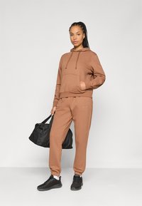 Brown fleece hoodie with a front pocket and drawstrings, paired with matching joggers and black athletic shoes, holding a black duffel bag.
