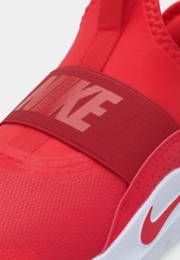 Scarpa sportiva rossa con tomaia liscia e texture, una larga fascia elastica con il marchio "NIKE" e una suola in gomma bianca con il logo swoosh.