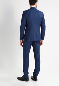 Abito blu con una vestibilità su misura, dotato di uno spacco singolo sul retro. Tessuto liscio, pantaloni con frontale piatto, abbinati a scarpe nere lucide.