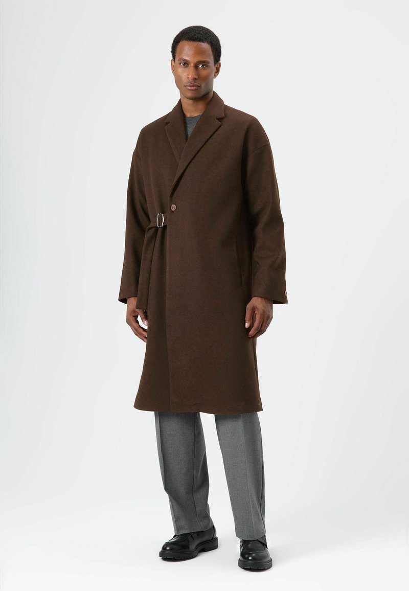 Bruine jas van een wolmengsel met een enkele knoopsluiting en een ceintuurdetail aan de zijkant, gecombineerd met grijze broek en zwarte schoenen. Soepele textuur, getailleerde pasvorm.