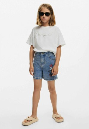 Camiseta blanca de manga corta con "Desigual" en letras brillantes, pantalones cortos de mezclilla azules con bordado de Minnie Mouse, chanclas beige, gafas de sol negras.