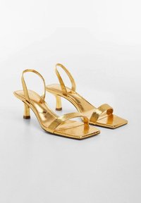 Gold heeled sandals with a square toe, featuring thin straps and a textured, glossy finish. The heel is a short, tapered design.