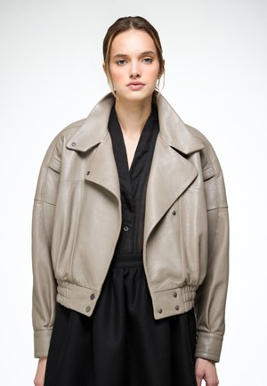 Mladá žena v volné taupe kožené bundě přes černé šaty s výstřihem do V, stojící na jednobarevném bílém pozadí.