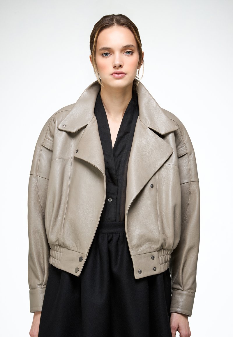 Jeune femme portant une veste en cuir taupe ample sur une robe noire à col en V, posant devant un fond blanc uni.