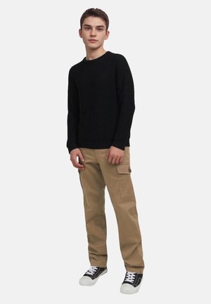 Schwarzer gestrickter Pullover, beige Cargo-Hose mit Seitentaschen und schwarze Sneakers mit weißen Sohlen. Einfaches, lässiges Outfit an einer stehenden Figur.