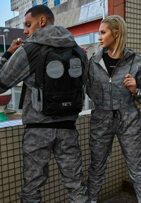 Tute grigi a motivo camouflage con polsini elasticizzati e cordini regolabili, abbinati a uno zaino nero con tasche circolari grigie e cerniere.