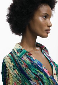 Cămașă multicoloră cu texturi unduitoare în verde, roz și albastru, cu guler și accente cu nasturi. Model detaliat al țesăturii vizibil.