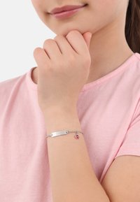 Una pulsera de plata presenta una placa de identificación plana y un pequeño colgante en forma de corazón rojo, descansando sobre una muñeca con una camiseta de color rosa claro de fondo.
