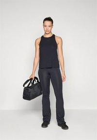 Μαύρο αθλητικό τοπ χωρίς μανίκια και εφαρμοστά παντελόνια, και τα δύο από λείο ύφασμα. Η μοντέλο κρατά μια μαύρη τσάντα ταξιδιού με διπλές λαβές στην κορυφή.