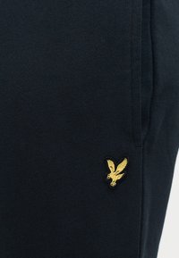 Mørk marineblå joggebukser med en glatt tekstur, med en gullbrodert ørn-logo på venstre side. Enkel design, elastisk midjebånd.