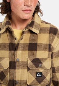Braun-gelb kariertes Fleece-Hemdjackett mit zwei Brusttaschen, Knopfverschluss und einem Logopatch auf der linken Tasche. Weiche Textur.