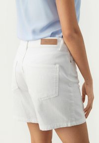 Valkoiset denimit sortit, joissa on takataskut ja ruskea nahkaleima vyötäröllä. Kankaalla on sileä rakenne ja se on istuva.