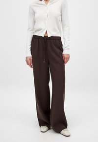 Braune, weite Hosen mit einem Kordelzugbund und weicher, glatter Textur, kombiniert mit einem weißen, knöpfbaren Cardigan. Schlichtes Design, keine Muster.