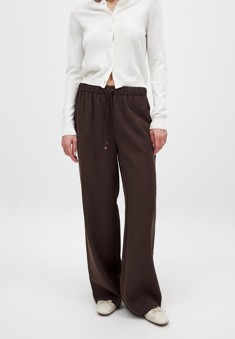 Braune, weite Hosen mit einem Kordelzugbund und weicher, glatter Textur, kombiniert mit einem weißen, knöpfbaren Cardigan. Schlichtes Design, keine Muster.