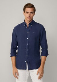 Camicia di lino blu navy con colletto buttons-down, maniche lunghe arrotolate e piccolo logo ricamato sul petto sinistro. Tessuto testurizzato.