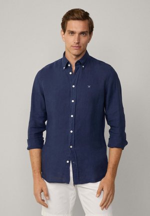 Navy blue linen shirt with button-down collar, long sleeves rolled up, and small embroidered logo on the left chest. Textured fabric.