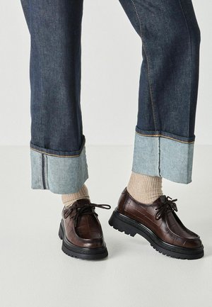 Fødder iført mørkebrune teksturerede læder snørestøvler, beige ribstrikkede sokker og opsmøgede mørkeblå jeans med en bred lys denimkant.