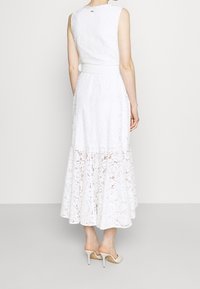 Kvinna som bär en ärmlös vit spetsmidi-klänning med bälte, stående bakifrån, tillsammans med vita högklackade mule-skor på en enkel bakgrund.